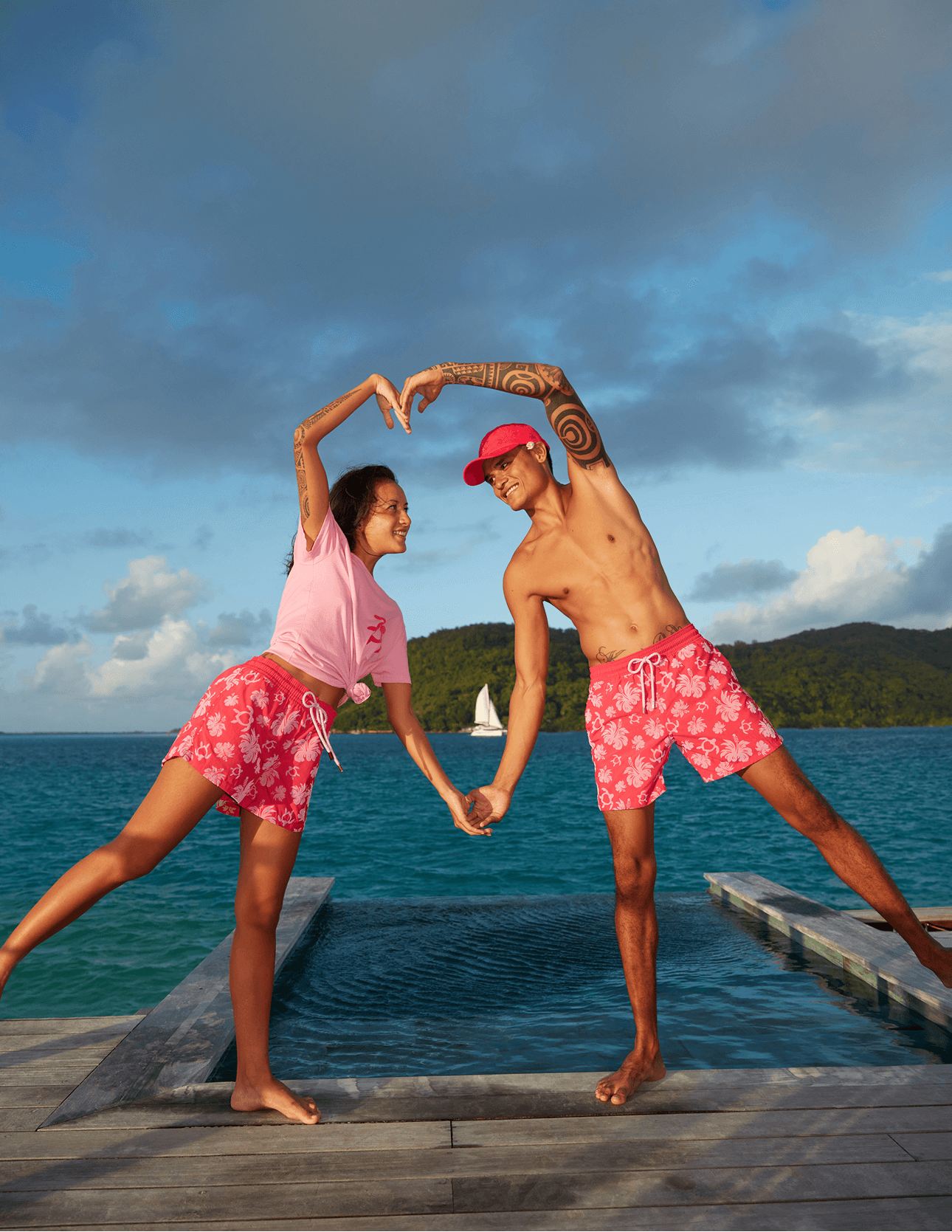 A man and a womand doing a heart with their arms, in front of the sea. They are wearing turtle heart pattern swimwear